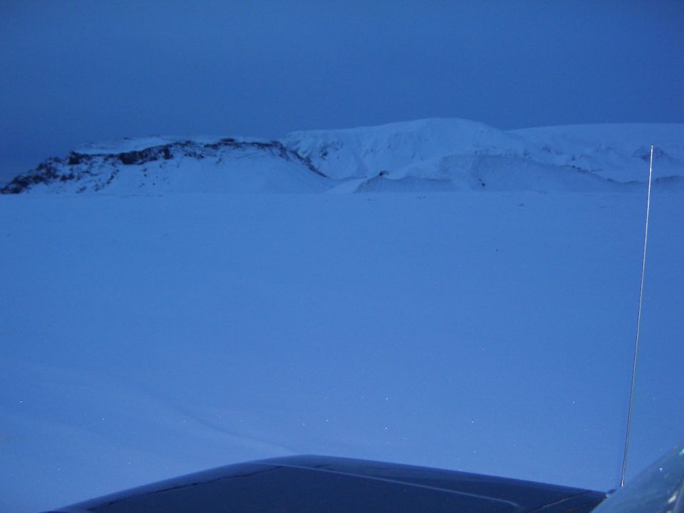Snowy tundra at dusk seen over a dark vehicle hood with antenna