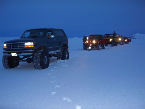 1996 Ford Bronco - A convoy of off-road vehicles, including a 1996 Ford Bronco, travels across a snowy landscape under a twilight sky.