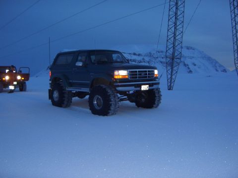 1996 Ford Bronco - A dark green 1996 Ford Bronco with oversized tires drives through a snowy landscape near power lines.