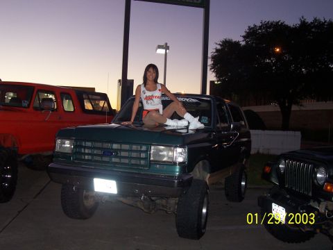 1989 Ford Bronco - A woman poses on the hood of a lifted green Ford Bronco, with other vehicles in the background.