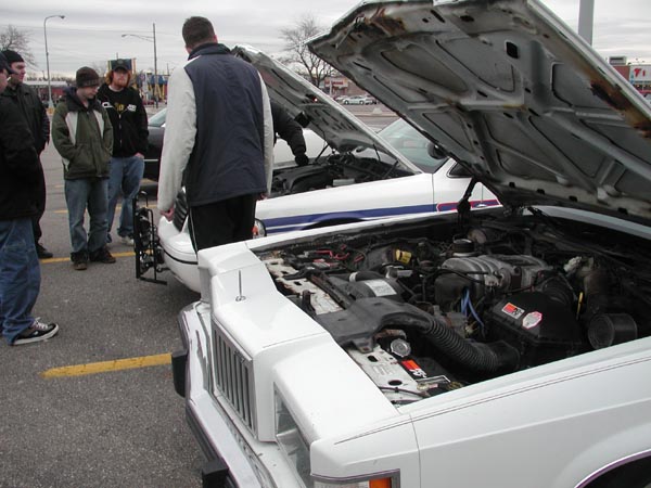 Two white sedans with hoods open, showing engines. People stand around the cars