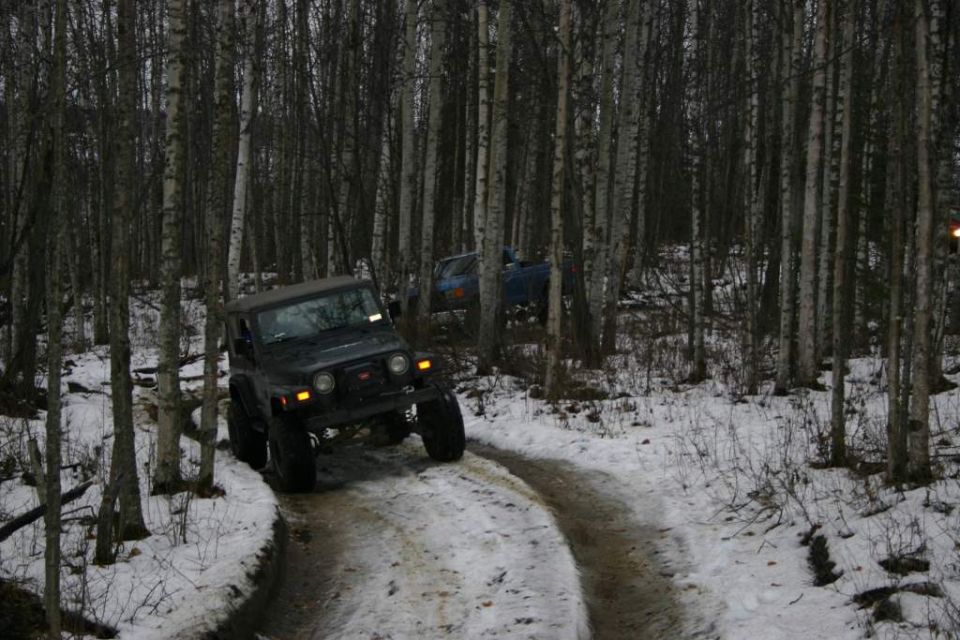 2003 Jeep TJ navigating a snowy path through a birch forest; another blue vehicle is visible deeper in the woods