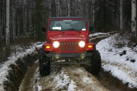 2003 Jeep TJ - A red 2003 Jeep TJ navigates a challenging muddy and snowy path in a wooded area.