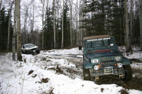 2003 Jeep TJ - Two off-road vehicles are stuck in a snowy and muddy forest trail, with one vehicle winching the other.