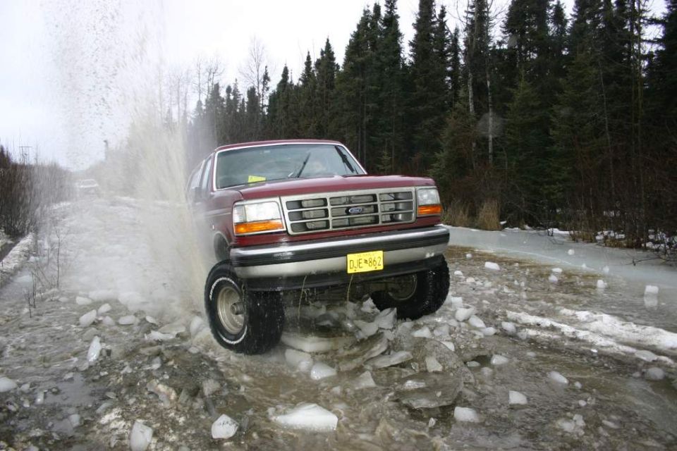 Red Ford Bronco splashing through icy water on a dirt road; another vehicle visible in the distance