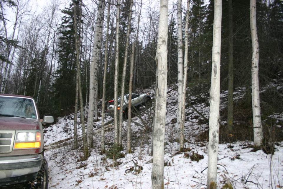 Maroon truck in foreground, white SUV climbing a steep, snowy hill in woods