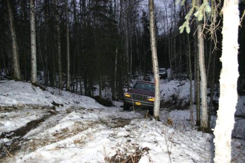2003 Jeep TJ - A red pickup truck navigates a snow-covered trail in a wooded area, with a second vehicle visible further up the path.