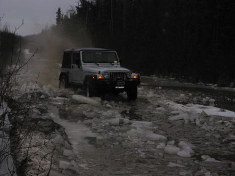 1993 Ford Bronco - A silver Jeep Wrangler navigates a treacherous icy and slushy road through a wooded area.