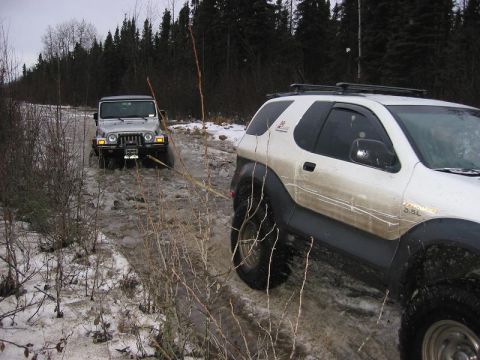 1993 Ford Bronco - Two SUVs navigate a challenging muddy and snowy trail through a forest.