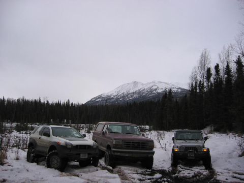 1993 Ford Bronco - Three SUVs, including a 1993 Ford Bronco, are parked in a snowy, wooded area with a mountain in the background.