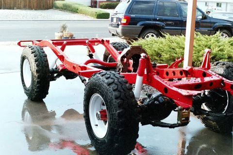 1976 Toyota Land Cruiser - A red vehicle frame with large off-road tires sits on a wet surface, with a blue SUV parked behind it.