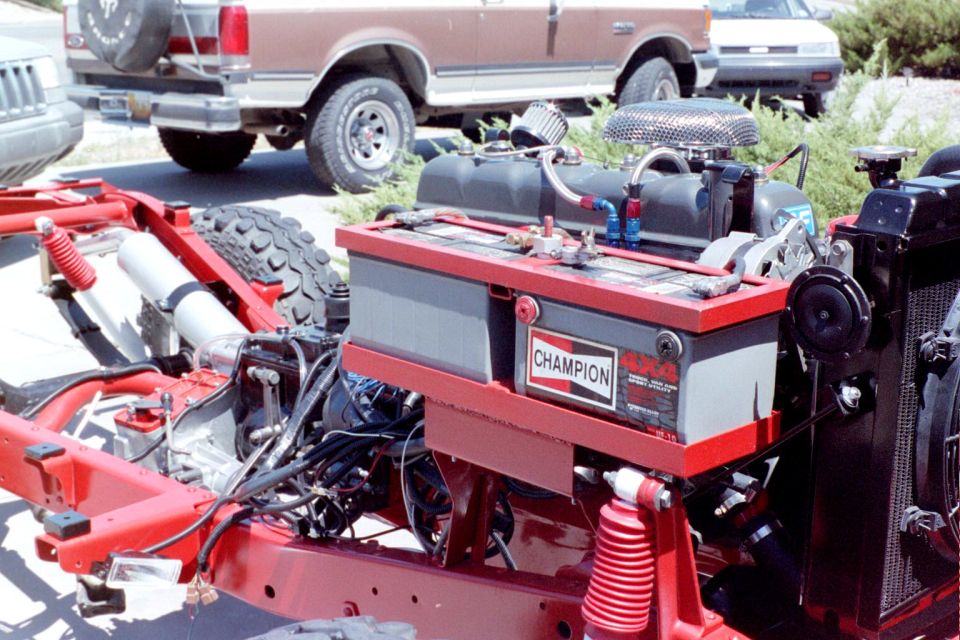 Red rolling chassis with engine, battery box and Champion 4x4 sticker; two-tone pickup in background