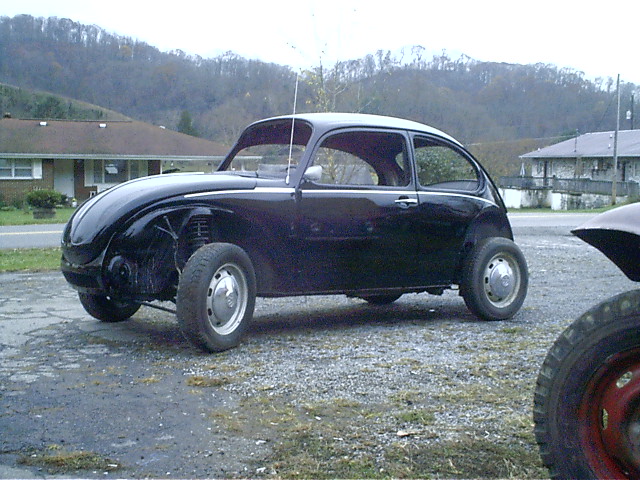 Custom black chopped-roof beetle-shaped car on steel wheels parked on gravel by rural houses