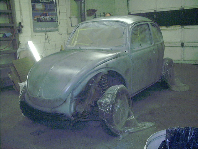 Primer-gray Beetle-style body shell mid-restoration on stands in a garage, wheels wrapped in plastic