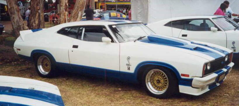 White Ford Falcon coupe with blue stripes and hood scoop, parked next to another white car