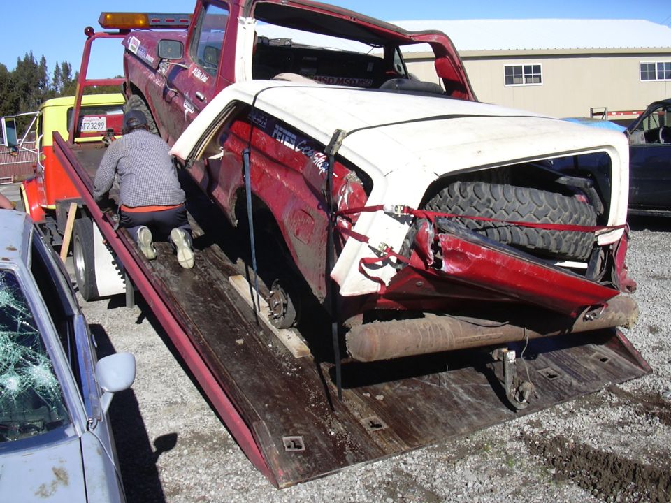 red and white pickup truck with significant rear damage being loaded onto a tow truck, spare tire visible in the back