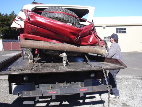1978 Ford Bronco - A heavily damaged red 1978 Ford Bronco is secured to a tow truck, showing extensive rear-end collision damage.