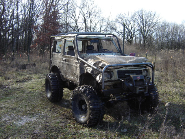 Modified Suzuki Samurai with large tires and roll cage parked in a grassy, wooded area