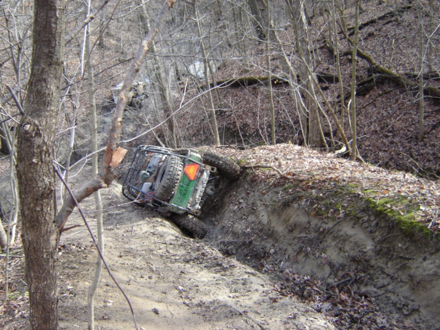 Green off-road vehicle tilted on a muddy bank with another vehicle behind it in the woods