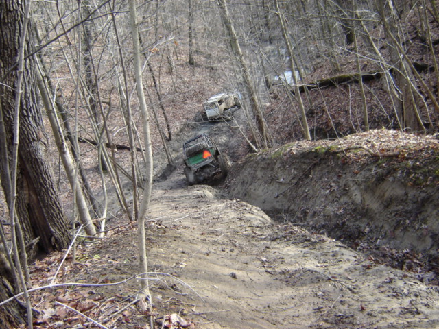 Two off-road vehicles navigate a muddy, wooded incline