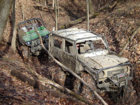 1992 Ford Bronco - Two off-road vehicles navigate a muddy, wooded trail. The lead vehicle is a white Suzuki Samurai, followed by a green utility vehicle.