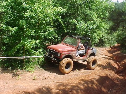 1978 Ford Bronco - A red off-road vehicle navigates a dusty trail through a wooded area.
