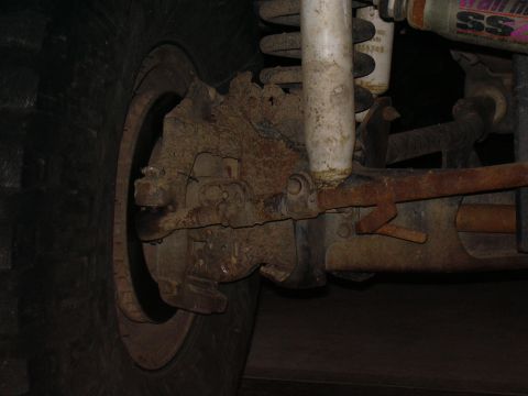 1991 Ford Bronco - Close-up underside view of a solid front axle showing the differential/knuckle area caked in mud, with a white shock absorber, coil spring,…