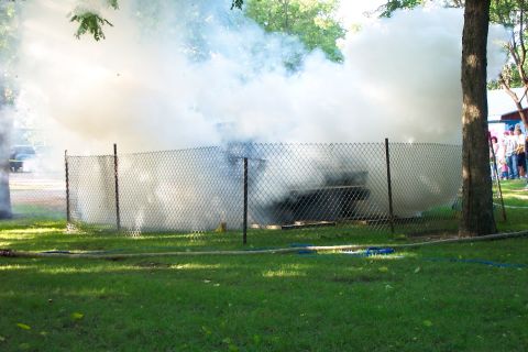 1977 Ford F-150 - A vehicle is engulfed in smoke behind a chain-link fence on a sunny day.