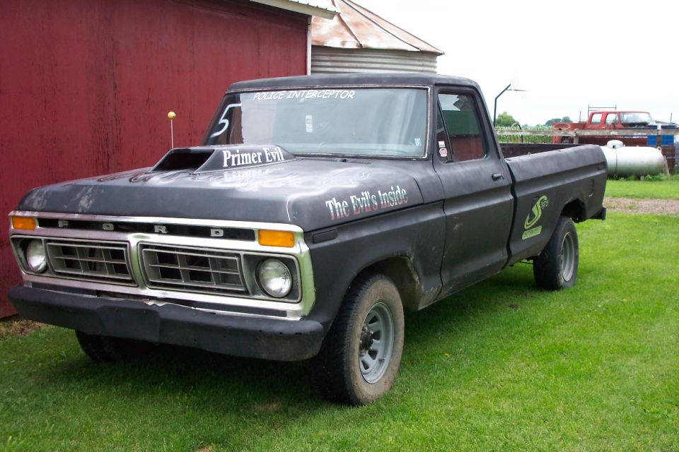Primer-black 1970s Ford pickup with hood scoop and Primer Evil lettering by a red barn