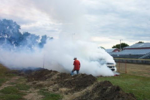 1977 Ford F-150 - A truck is engulfed in smoke, with a person standing in the foreground. A stadium with bleachers is visible in the background.