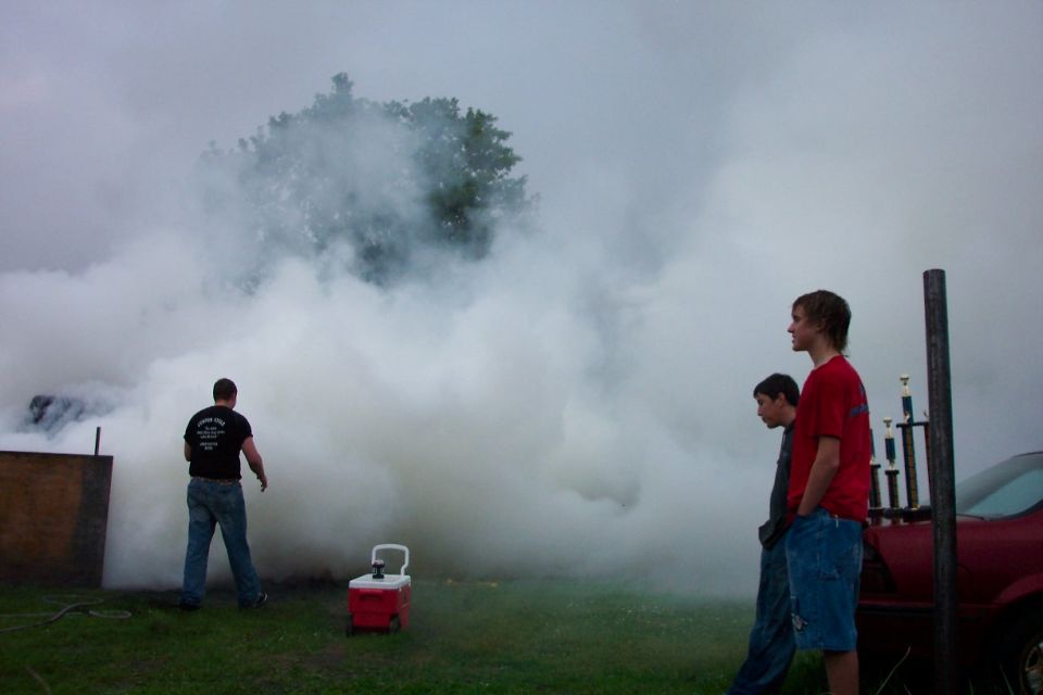 Thick white tire smoke from a burnout with spectators and trophies at a grassy lot.