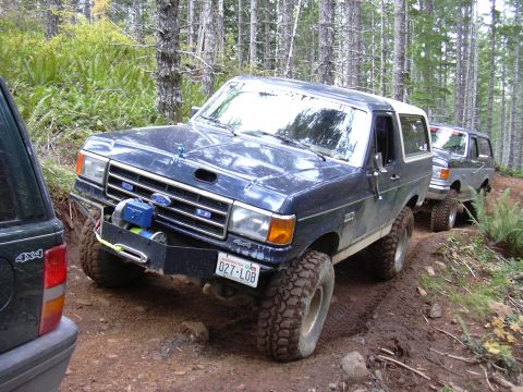 1991 Ford Bronco - Two Ford Broncos navigate a muddy off-road trail through a forest.