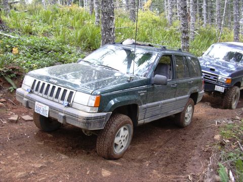 1991 Ford Bronco - A green Jeep Grand Cherokee drives on a muddy forest road, with another vehicle behind it.