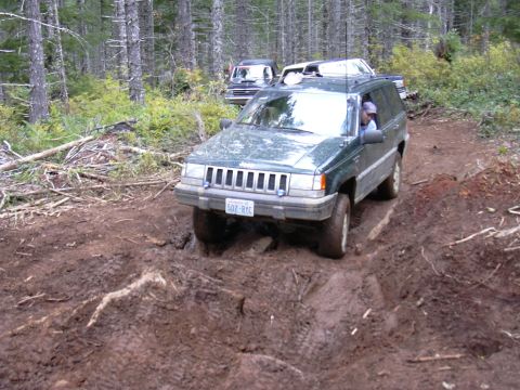 1991 Ford Bronco - A green Jeep Grand Cherokee tackles a challenging muddy trail, with another vehicle following in the background.