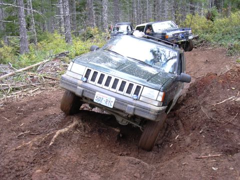 1991 Ford Bronco - Multiple SUVs tackle a challenging muddy path in a wooded area.
