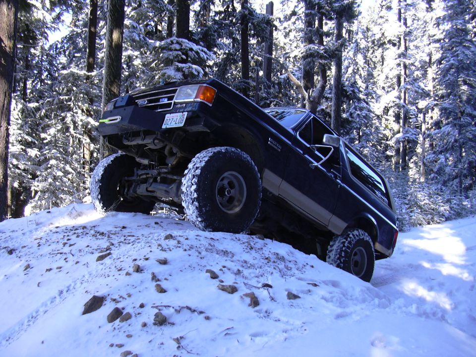 Black Ford Bronco climbing a snowy hill in a forest, front-left quarter view