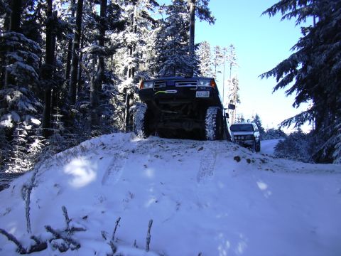 1991 Ford Bronco - A dark 1991 Ford Bronco navigates a snow-covered incline, with another vehicle visible behind it on the trail.