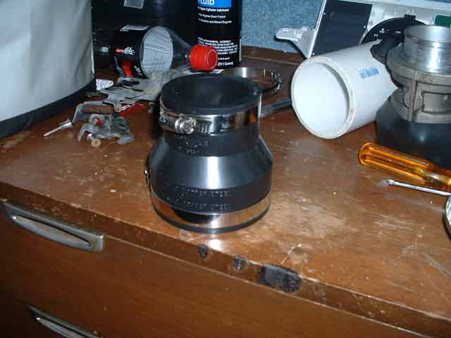 Close-up of a black rubber automotive intake hose with metal clamps, sitting on a wooden surface next to a white pipe and a screwdriver