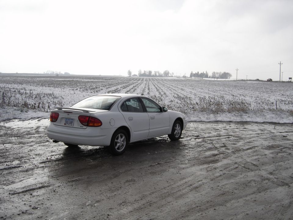 White Oldsmobile Alero sedan with rear spoiler parked by a snowy cornfield