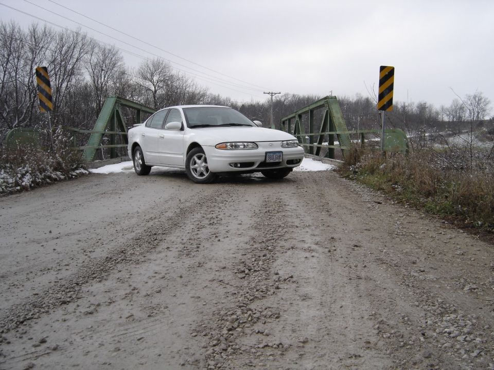 White Oldsmobile Alero sedan on a snowy gravel road by a green truss bridge