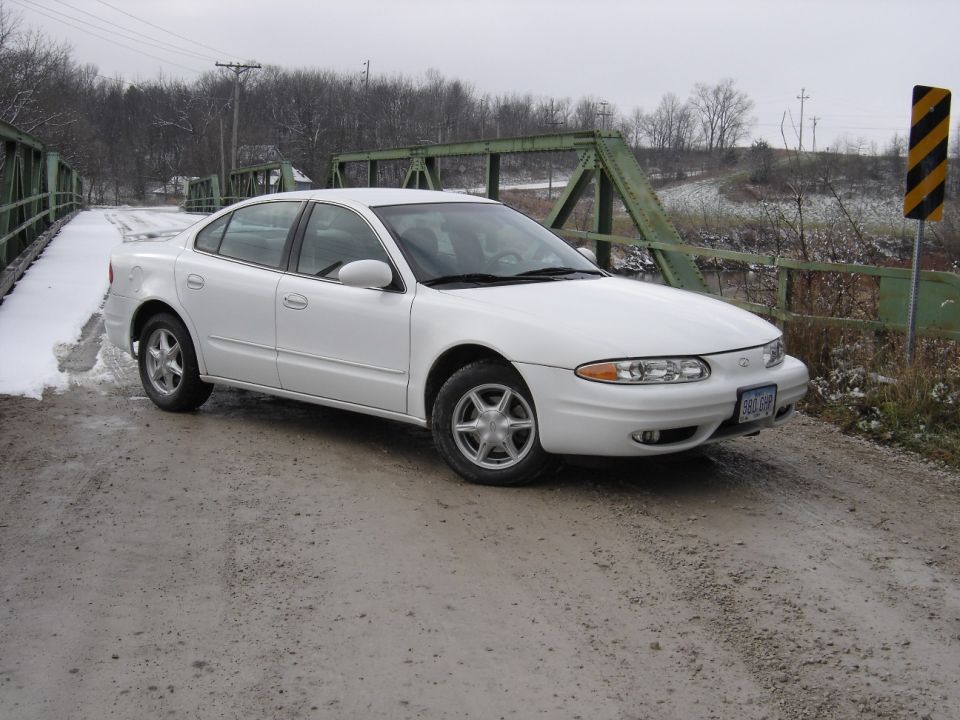 White Oldsmobile Alero sedan parked on gravel by a green truss bridge in winter.