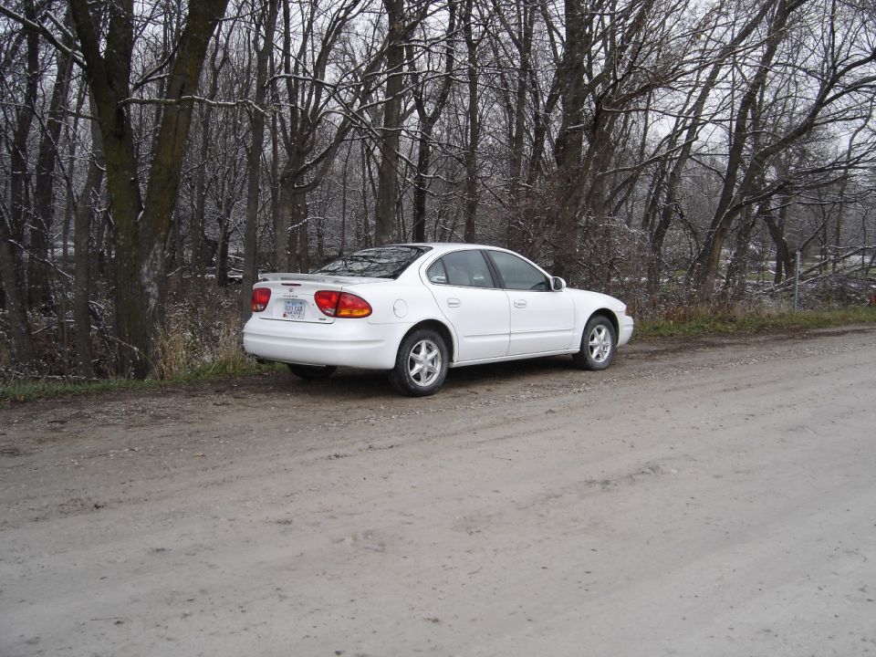 White sedan parked on dirt road by leafless trees, rear three-quarter view