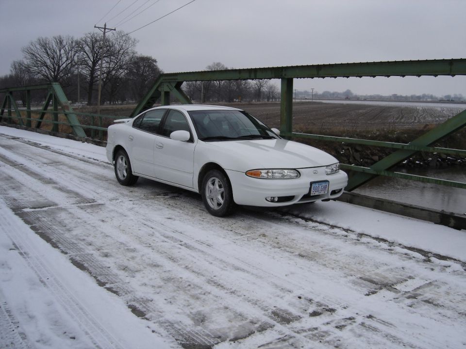 White Oldsmobile Alero sedan on a snowy steel truss bridge in winter, front three-quarter view