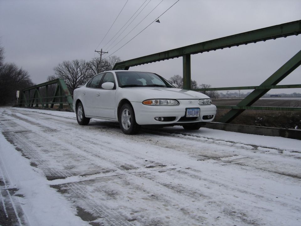 White Oldsmobile Alero sedan on a snowy green truss bridge, 3/4 front view