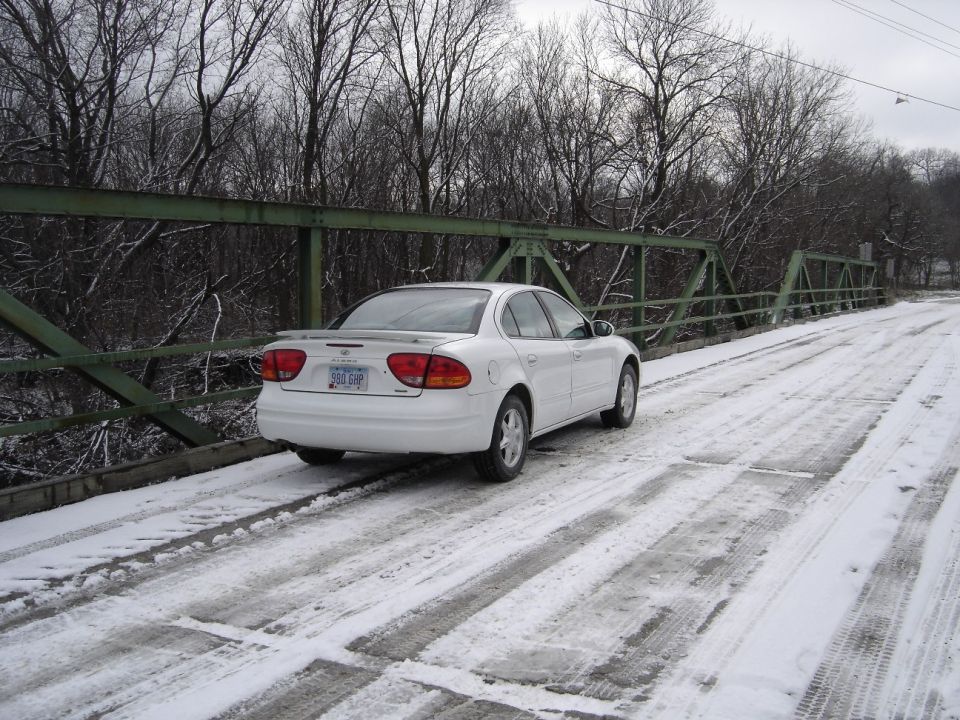 White Oldsmobile Alero sedan parked on a snowy green steel truss bridge among bare trees