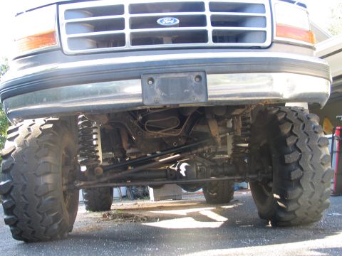 1992 Ford Bronco - Worm's-eye view under the front of a lifted Ford truck with a chrome grille and blue Ford oval, showing a solid front axle with centered di…