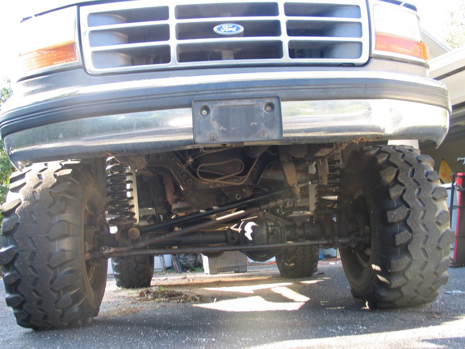 Low front view under a lifted Ford showing solid front axle, coil springs, and big tires