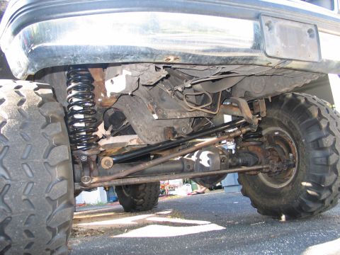1992 Ford Bronco - Low-angle underbody view of a lifted 4x4 showing the front solid axle, coil spring, steering linkage (tie rod and drag link), and a black s…