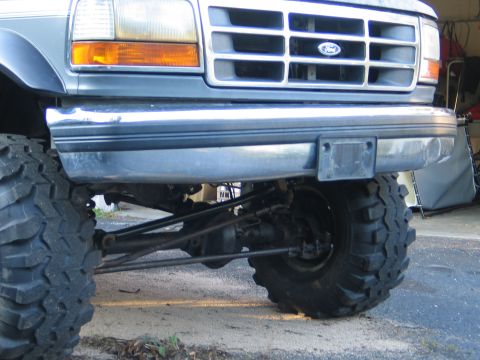 1992 Ford Bronco - Low front view of a lifted 1990s Ford Bronco in dark blue/gray with a chrome bumper and Ford grille badge, showing large mud-terrain tires…