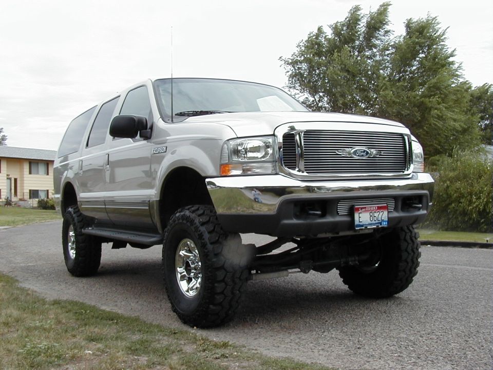 Lifted white Ford Excursion SUV with big off-road tires and billet grille, front three-quarter view on a street